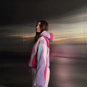 May include: A color-block hoodie in shades of pink, purple, and blue. The hoodie features a drawstring and a text detail on the sleeve. The model is wearing matching pink sweatpants. The background is a metallic wall.