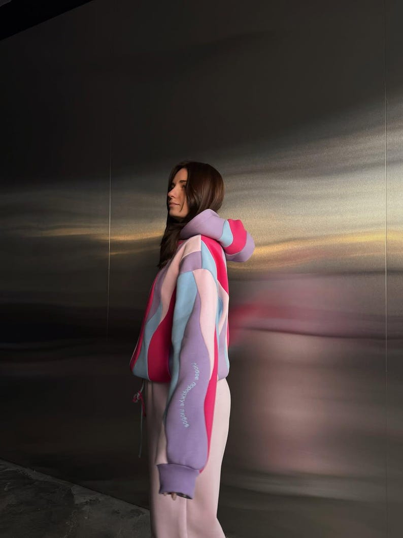 May include: A color-block hoodie in shades of pink, purple, and blue. The hoodie features a drawstring and a text detail on the sleeve. The model is wearing matching pink sweatpants. The background is a metallic wall.