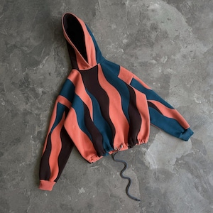 Puede incluir: Una sudadera con capucha recortada con un patrón ondulado único en coral, verde azulado y marrón. La sudadera tiene un cordón en la cintura y una capucha. Las mangas son largas y los puños son de color coral. El diseño es moderno y llamativo.
