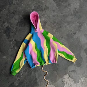 Op de afbeelding: Een kleurrijke hoodie met een golvend patroon in geel, groen, blauw en roze. De binnenkant van de capuchon is roze. Het heeft een trekkoord in de taille en groene en gele manchetten. Een stijlvol kledingstuk.