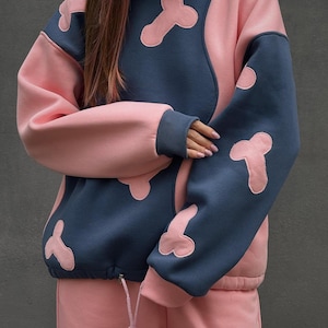 May include: A two-tone hoodie and matching sweatpants set. The hoodie is navy blue with pink accents and bone-shaped designs. The sweatpants are pink. The drawstring on the hoodie is visible.
