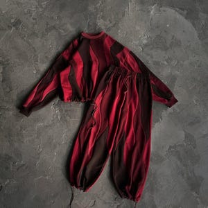 May include: A two-piece maroon and dark brown outfit. The top is a long-sleeved cropped sweatshirt with vertical wavy panels. The matching pants have a drawstring waist and are gathered at the ankles. The outfit is displayed on a gray surface.