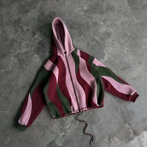 Pode incluir: Um casaco com capuz e fecho de correr apresentando um padrão ondulado multicolorido em tons de rosa claro, bordeaux escuro e verde azeitona. A peça inclui um fecho frontal completo, um cordão na bainha e mangas compridas com punhos.