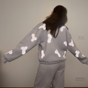 Puede incluir: Un conjunto de sudadera de manga larga y pantalones de chándal de color gris claro. La sudadera presenta un patrón repetitivo de estampados gráficos blancos con forma fálica distribuidos por la espalda, las mangas y los hombros.