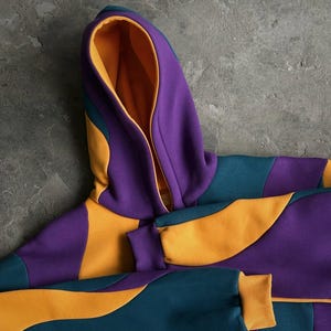 May include: A color-blocked hoodie in shades of purple, teal, and yellow. The hood is lined with yellow fabric, and the cuffs are yellow. The hoodie has a full zipper and a modern, geometric design.