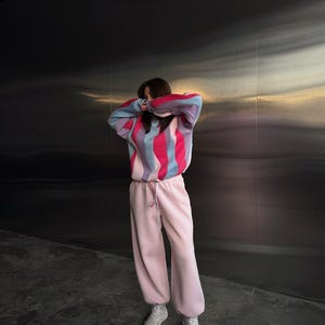 May include: A person wearing a colorful striped sweater with pink, blue, and red vertical stripes. The person is also wearing light pink sweatpants and white sneakers. The background is a dark, textured wall.
