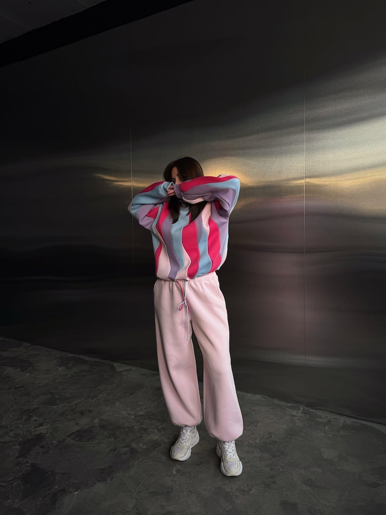 May include: A person wearing a colorful striped sweater with pink, blue, and red vertical stripes. The person is also wearing light pink sweatpants and white sneakers. The background is a dark, textured wall.