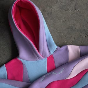 May include: A colorful hoodie in shades of pink, purple, and blue. The hoodie features a layered design with a contrasting pink interior lining the hood. The sleeve has embroidered text. The garment is made of a soft material.
