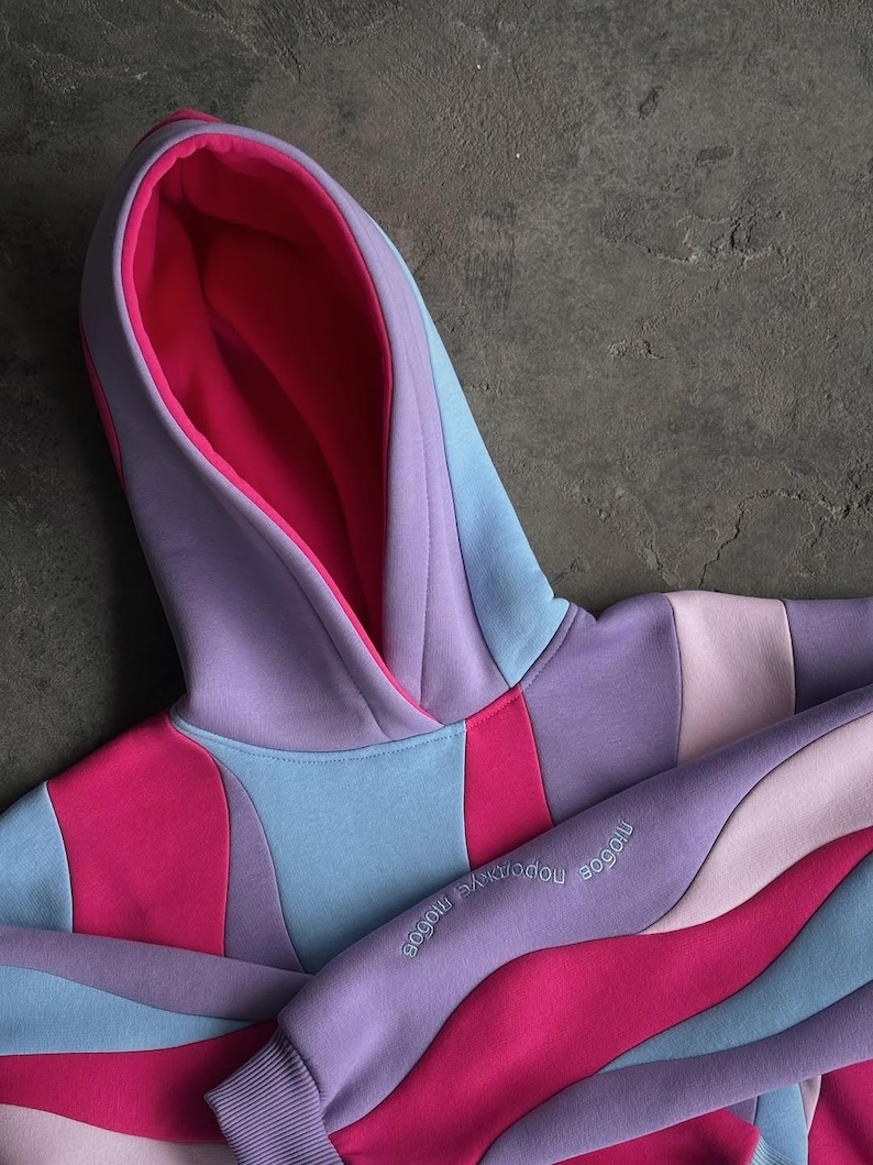 May include: A colorful hoodie in shades of pink, purple, and blue. The hoodie features a layered design with a contrasting pink interior lining the hood. The sleeve has embroidered text. The garment is made of a soft material.
