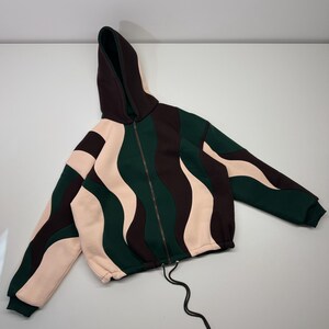 May include: A zip-up hooded sweatshirt featuring a wavy, colour-blocked pattern in dark brown, forest green, and pale pink. The garment includes a full-length front zip, a drawstring at the hem, and long sleeves with ribbed cuffs.