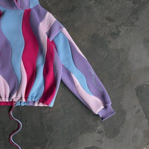 May include: Cropped hoodie with a wavy pattern in shades of purple, pink, and blue. The drawstring is two-toned, and the sleeve features embroidered text. The hoodie has a hood and a ribbed cuff.