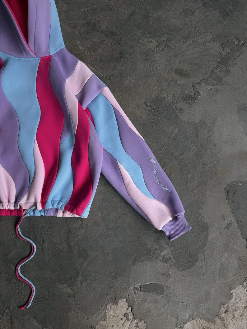 May include: Cropped hoodie with a wavy pattern in shades of purple, pink, and blue. The drawstring is two-toned, and the sleeve features embroidered text. The hoodie has a hood and a ribbed cuff.