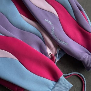 May include: A multi-colored sweatshirt with a unique wavy design. The sweatshirt features shades of pink, purple, and blue. The sleeves have ribbed cuffs and drawstrings. The text "любовь порождает любовь" is embroidered on the sleeve.