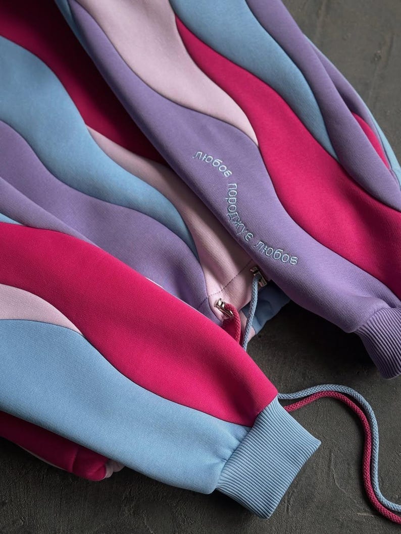 May include: A multi-colored sweatshirt with a unique wavy design. The sweatshirt features shades of pink, purple, and blue. The sleeves have ribbed cuffs and drawstrings. The text "любовь порождает любовь" is embroidered on the sleeve.