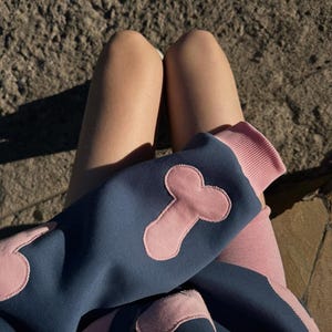 Puede incluir: Una sudadera azul marino con detalles en rosa y un diseño abstracto en rosa. La sudadera tiene un puño acanalado rosa. La persona lleva leggings rosas. El fondo es una superficie de piedra.