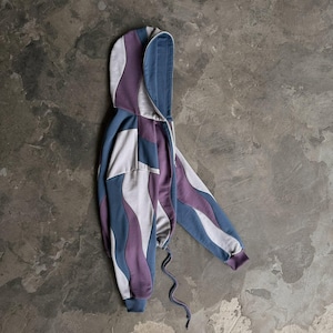 May include: A hooded zip-up sweatshirt in a unique wavy pattern. The design incorporates shades of blue, purple, and gray. The sweatshirt has a drawstring at the waist and ribbed cuffs. The garment is laid flat on a textured gray surface.