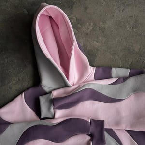 May include: A pink, gray, and purple hoodie with a wave pattern design. The hoodie features a lined hood and ribbed cuffs. The color scheme includes shades of pink, gray, and purple, creating a modern and stylish look. The hoodie is made of a soft material.