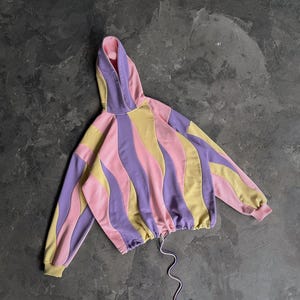 May include: A cropped hoodie in a pastel color block design. The hoodie features wavy vertical stripes in pink, yellow, and purple. It has a drawstring at the waist and a hood. The sleeves and hood lining are pink.