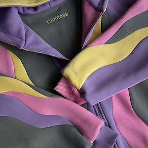 May include: A zip-up hoodie in a unique design, featuring wavy panels of gray, purple, pink, and yellow. The cuffs and hood lining are a solid color, and the brand name "roomova" is embroidered on the inside collar.