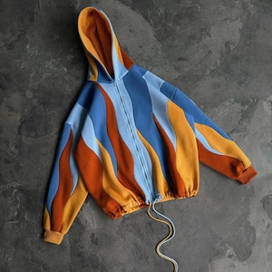 Könnte beinhalten: Ein Kapuzen-Sweatshirt mit Reißverschluss in einem einzigartigen Design. Die Jacke hat ein Colorblock-Muster mit Blautönen, Orange und Gelb. Der Kordelzug an der Taille und die Manschetten sind orange. Die Innenseite der Kapuze ist orange.