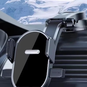 May include: A black and silver car phone holder with a glossy finish. The holder has adjustable arms and a secure grip to hold a phone. The background shows a snowy mountain range.