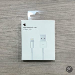 Könnte beinhalten: Weißes Lightning-auf-USB-Kabel, 1 Meter lang, in einer weißen Box mit dem Apple-Logo. Die Box hat einen transparenten Kunststoffaufhänger. Das Kabel hat einen Lightning-Anschluss und einen USB-Anschluss.
