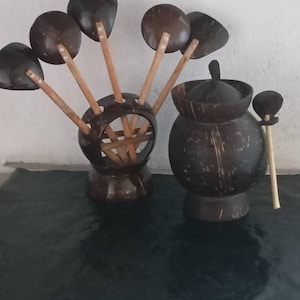 Puede incluir: Un juego de utensilios de cocina hechos a mano y un recipiente con tapa hechos de cáscaras de coco. Los utensilios incluyen un soporte con múltiples cucharas y una olla pequeña con tapa. Los artículos son de color marrón oscuro y descansan sobre una superficie oscura.