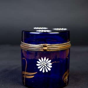 Könnte beinhalten: Ein zylindrisches Glasgefäß aus kobaltblauem Glas mit einem aufklappbaren Metalldeckel. Die Außenseite ist mit handgemalten weißen Gänseblümchenmotiven und goldenen Blattakzenten verziert. Der Deckel zeigt ebenfalls weiße Blumenmuster.