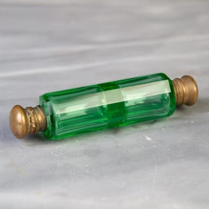 Può includere: Un flacone di profumo cilindrico in vetro verde con corpo sfaccettato e due tappi metallici alle estremità. Il vetro è traslucido e vivace, mentre i tappi presentano una finitura opaca e invecchiata. L'oggetto è allungato e simmetrico.