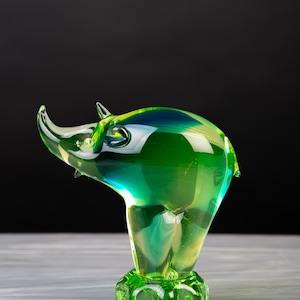 Può includere: Una statuetta di rinoceronte in vetro traslucido caratterizzata da un vibrante gradiente di tonalità verde smeraldo e giallo. La scultura presenta una superficie liscia e lucida con un corno ricurvo e poggia su una base circolare testurizzata in vetro verde.