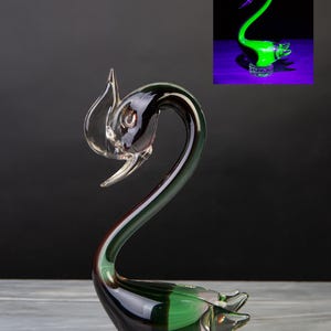 Può includere: Una statuetta di cigno in vetro con corpo verde scuro e nero su una base trasparente e testurizzata. Il cigno ha un collo lungo e curvo e una testa dettagliata. Un riquadro mostra lo stesso cigno che brilla di verde sotto luce UV.