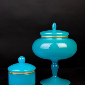 Pode incluir: Um pote de vidro turquesa brilhante com tampa e uma taça de vidro turquesa a condizer sobre um pedestal com tampa. Ambas as peças apresentam superfícies lisas e opacas, decoradas com finas bandas metálicas douradas e texturizadas.