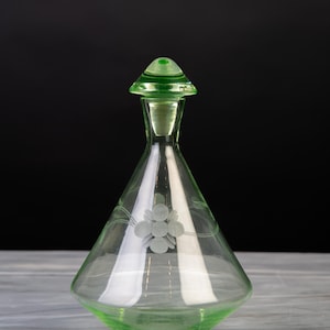 Op de afbeelding: Een lichtgroene glazen karaf met een conisch lichaam en een bijpassende puntige stop. Het glas is voorzien van een delicaat geëtst bloemmotief aan de voorzijde, met een centrale bloem en dunne, gebogen stelen die naar de zijkanten uitlopen.
