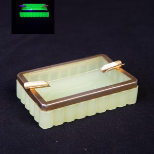 Può includere: Un posacenere rettangolare con base in vetro scanalato giallo pallido e bordo metallico color oro con due poggia-sigarette integrati. Un riquadro mostra lo stesso posacenere che brilla di un verde neon acceso sotto luce ultravioletta su sfondo nero.