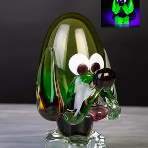 Puede incluir: Una figura de perro de cristal con cabeza de color verde oliva translúcido, orejas largas de color verde oscuro y una base de cristal transparente. El perro tiene grandes ojos circulares blancos con pupilas de color marrón oscuro y una nariz marrón oscuro. Un recuadro muestra el cristal brillando bajo luz ultravioleta.