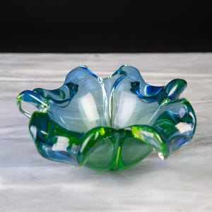 Op de afbeelding: Een decoratieve glazen schaal met een levendig kleurverloop van blauw en groen. De schaal heeft een opvallende, gegolfde, bloembladachtige rand met een doorschijnende afwerking die het licht opvangt en een gladde, gebogen, organische vorm creëert.