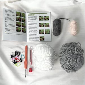 May include: A crochet kit with a booklet, yarn, and tools. The kit includes a crochet hook, yarn needles, stitch markers, and several skeins of yarn in white, gray, and pink. The booklet has instructions with images. The kit is on a white surface.