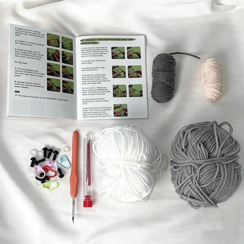 May include: A crochet kit with a booklet, yarn, and tools. The kit includes a crochet hook, yarn needles, stitch markers, and several skeins of yarn in white, gray, and pink. The booklet has instructions with images. The kit is on a white surface.