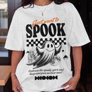 May include: A white short-sleeve t-shirt featuring a black graphic of a smiling ghost, a spider web, a moon, and spiders. The text reads: Just want to SPOOK, Embrace the spooky spirit and keep everyone on their toes!, and DECOCODE.