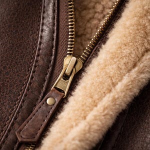 May include: Close-up of a brown leather jacket with a gold zipper and tan shearling lining. The leather has a textured appearance, and the zipper pull is made of brown leather.