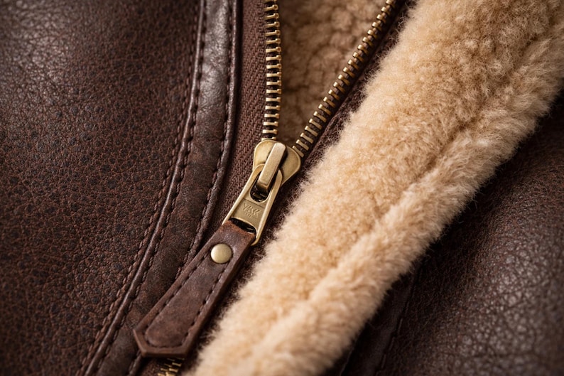 May include: Close-up of a brown leather jacket with a gold zipper and tan shearling lining. The leather has a textured appearance, and the zipper pull is made of brown leather.