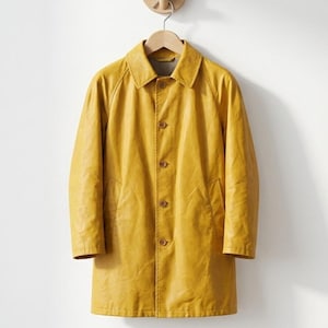 May include: A mustard yellow raincoat featuring a classic collar, long sleeves, and a front button closure with five visible buttons. The coat has a smooth, slightly textured finish and two side pockets, hanging on a wooden hanger against a wall.