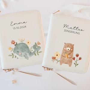 Könnte beinhalten: Zwei cremefarbene Reißverschluss-Geldbörsen mit verspielten Illustrationen. Eines zeigt einen grauen Wal mit floralen Akzenten und dem Namen "Emma" mit dem Datum "13.10.2024". Das andere zeigt einen braunen Bären mit einer Karotte und dem Namen "Matteo Zengerling".