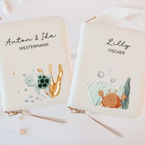 Könnte beinhalten: Zwei cremefarbene Reißverschlusstaschen mit aquatischen Illustrationen. Eine zeigt eine Schildkröte und den Text "Anton & Ida WESTERMANN", die andere eine Krabbe und den Text "Lilly FISCHER". Beide haben goldfarbene Reißverschlüsse und Handschlaufen.