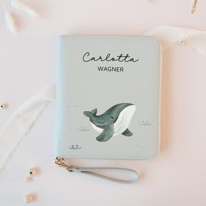 Könnte beinhalten: Hellblaues Portemonnaie mit Wal-Illustration und dem Aufdruck "Carlotta Wagner". Es hat einen Reißverschluss, eine Handschlaufe und einen goldfarbenen Reißverschluss.