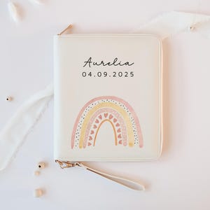 Könnte beinhalten: Cremefarbene Clutch mit Reißverschluss und einem Regenbogen-Design in Pastellrosa, Gelb und Pfirsich. Der Name "Aurelia" und das Datum "04.09.2025" sind auf der Vorderseite aufgedruckt. Mit Handschlaufe und goldfarbenem Reißverschluss.