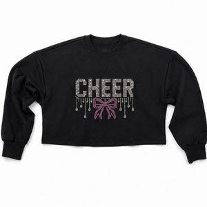 May include: A black long-sleeve cropped sweatshirt featuring the word CHEER in silver rhinestones. Below the text is a pink rhinestone bow graphic with silver rhinestone fringe accents hanging down from the letters and the bow design.