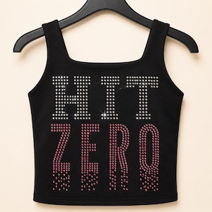 May include: A black sleeveless tank top featuring the word HIT in clear rhinestones and the word ZERO in pink rhinestones on the front. The garment has a square neckline and is displayed on a black plastic hanger against a light background.