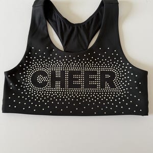 May include: Black sports bra with the word "CHEER" spelled out in silver rhinestones. The bra has a racerback design and is decorated with small rhinestones around the word.