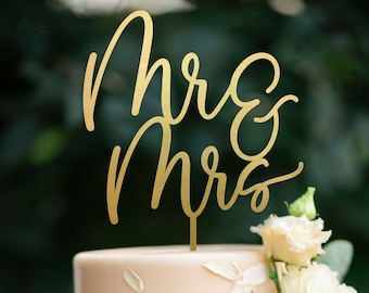May include: A two-tiered wedding cake with a light pink frosting and a wooden cake stand. The cake is decorated with white flowers and greenery. The cake topper says "Mr & Mrs".